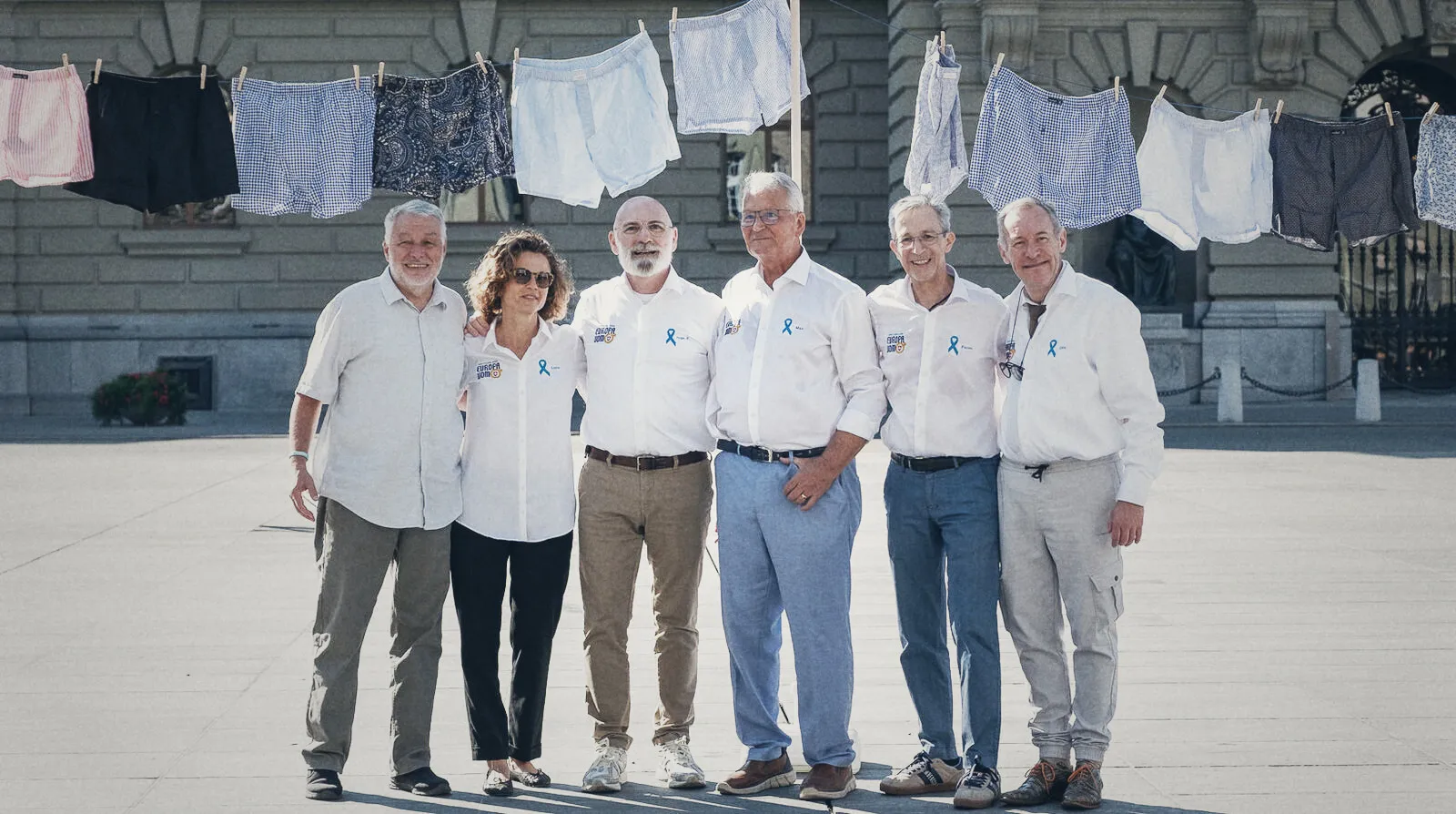 Vertreterinnen und Vertreter von Europa Uomo Schweiz in weissen Hemden mit blauer Schleife auf dem Bundesplatz in Bern, über ihnen eine Leine mit Boxershorts
